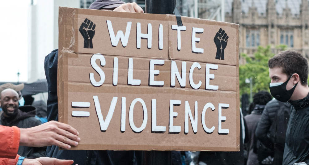 A handwritten sign at a protest reads 'white silence is violence' with two illustrations of black raised fists