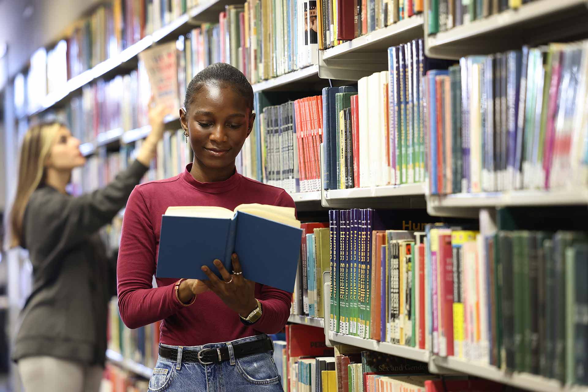 A student stands reading a book by book shelves in the library.