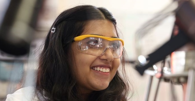 A close-up image of a woman with brown hair smiling and wearing protective goggles in a lab.