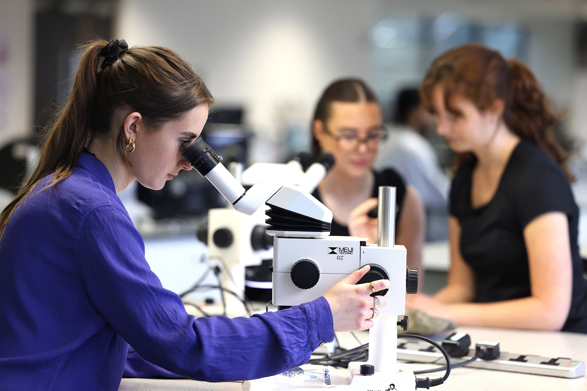 A female student in a royal blue lab coat looks into a microscope.