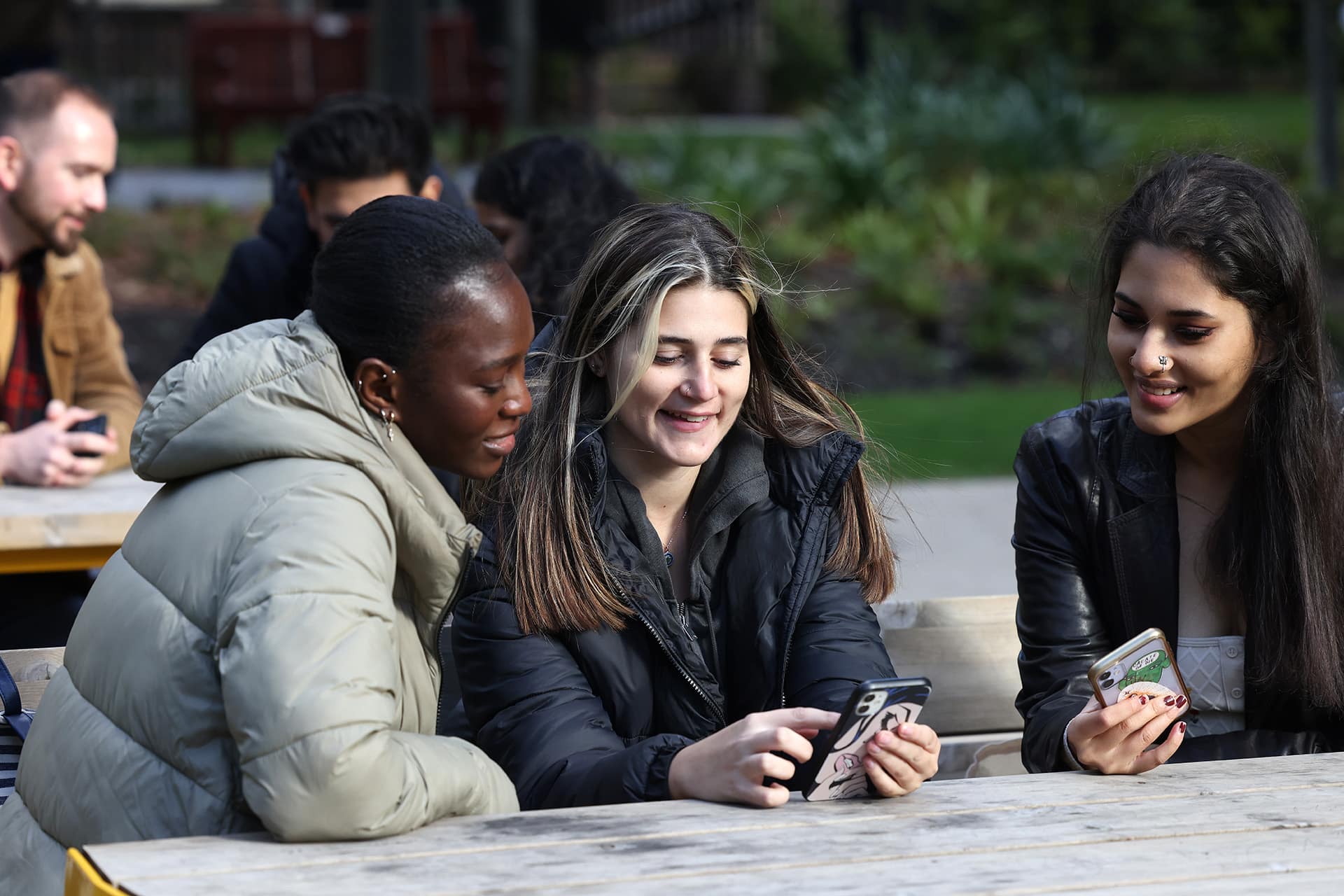 Three female students sit at an outside table on campus. They are smiling and looking at a phone together.