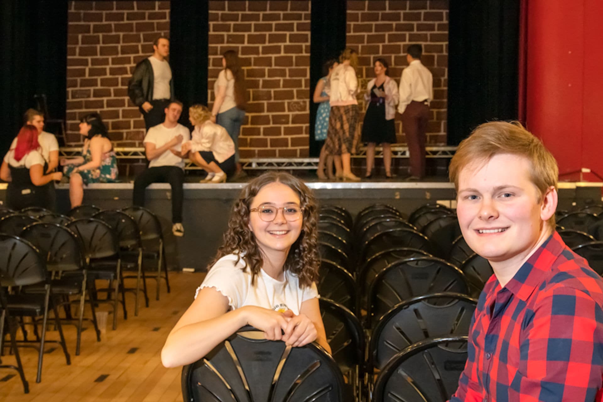 Students in theatre space smiling at camera with other students performing in background