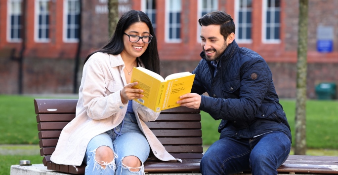 Two students read a book together while sat on a bench in the quad.