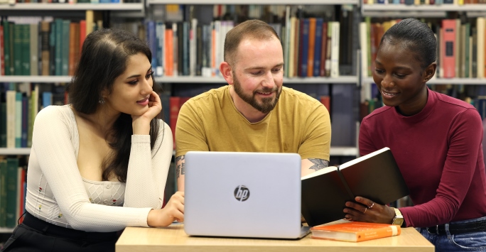 Three students consult a book and study a laptop while studying in the library.