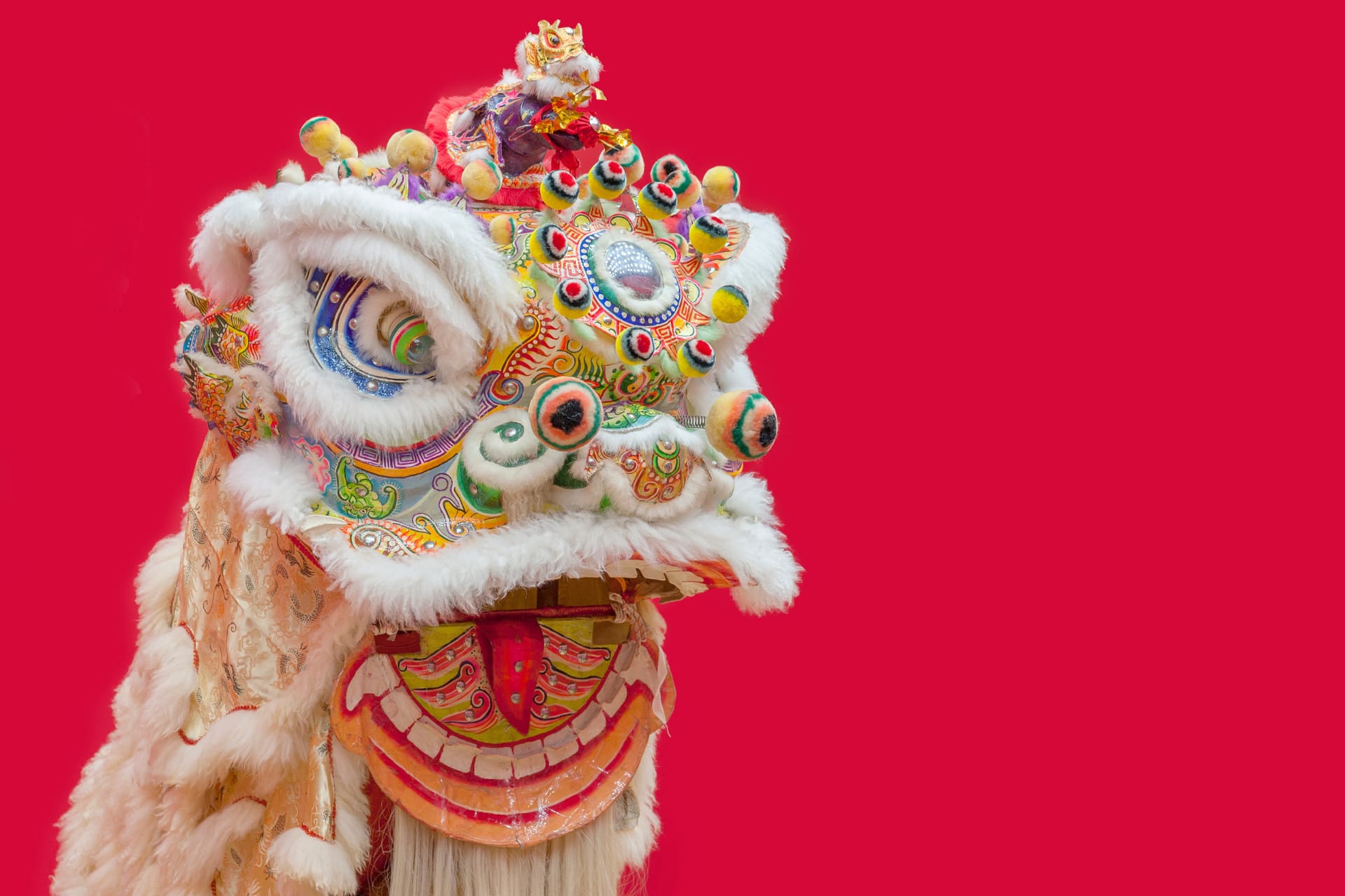 An ornate lion dance costume with white fun trim around the eyes and mouth on a bright red background.