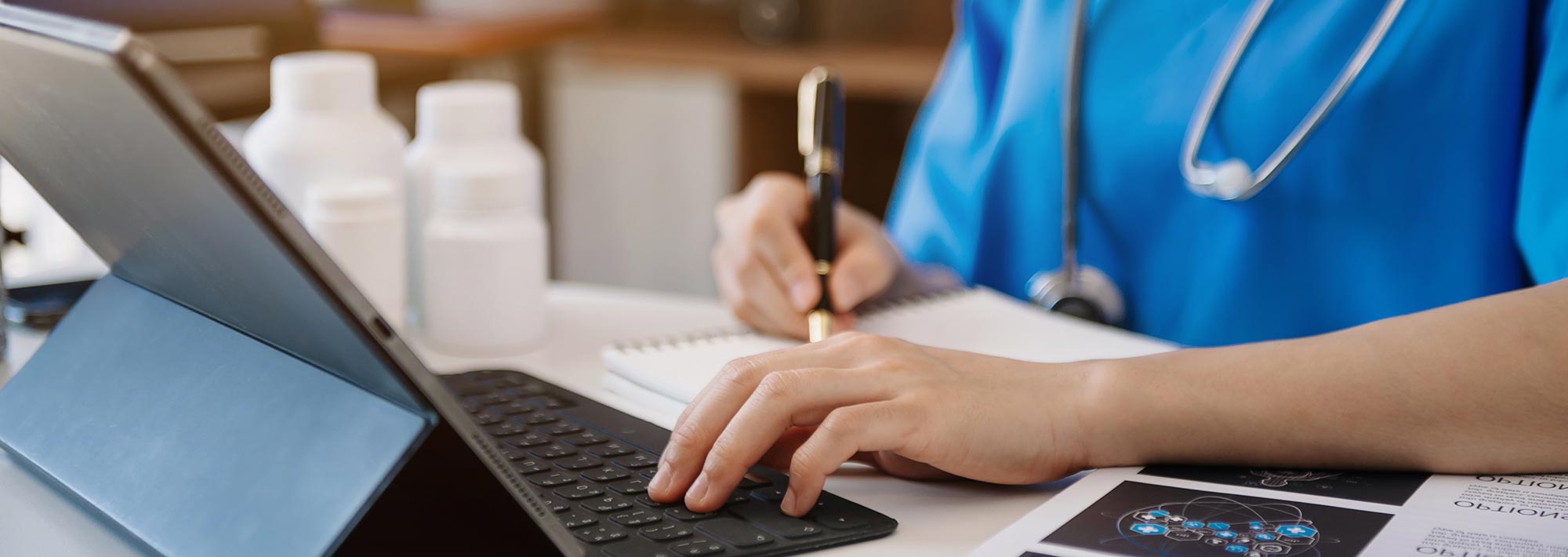 A professional with stethoscope makes notes while working on a tablet with keyboard.