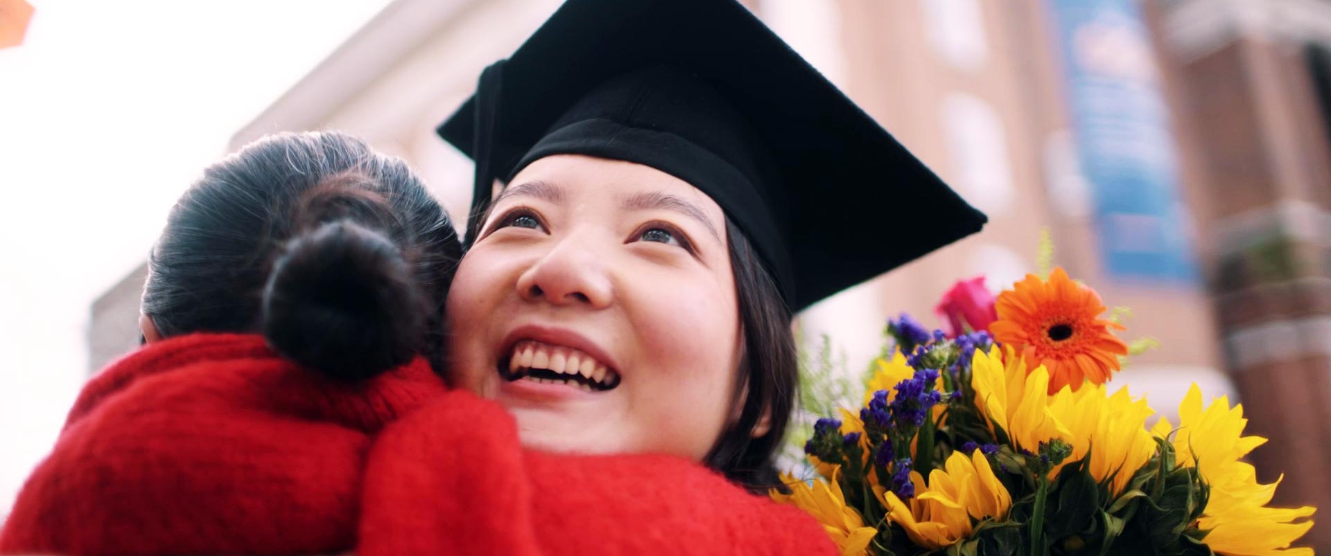A graduate embraces a loved one while holding a bouquet of flowers on their graduation day.