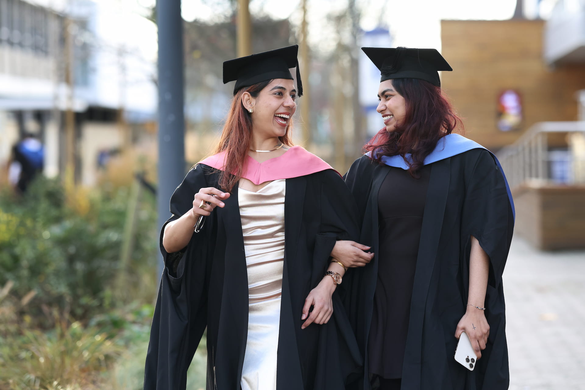 Two graduates in gowns and gaps smile at each other while walking