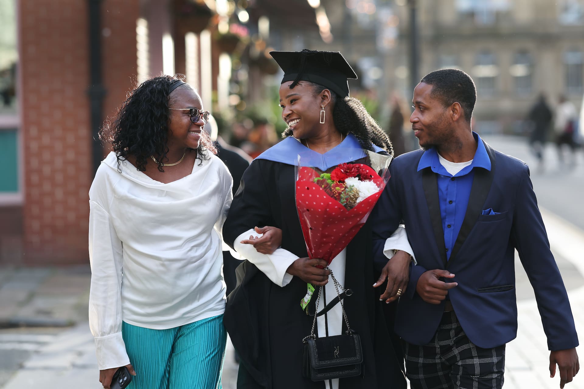 A graduate holding a bouquet of flowers walks arm in arm with her friends