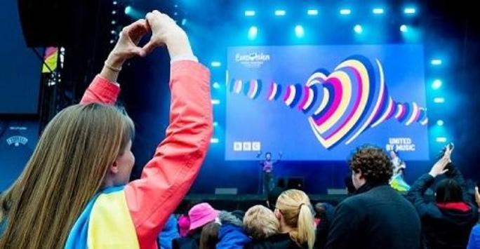 A crowd of people are watching a Eurovision 2023 event taking place on a large stage. In the foreground, a child is holding up their hands in a heart symbol.