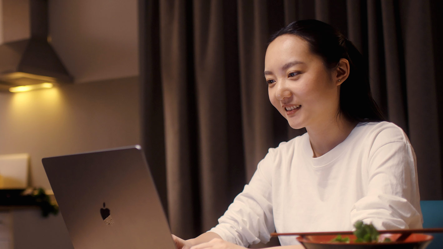 An image of a student sat at a desk looking at a laptop computer and smiling.