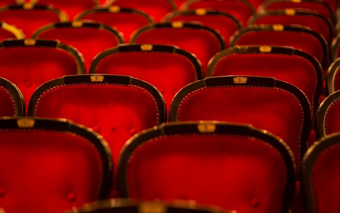 Two red theatre seats together in focus amongst several other red seats.