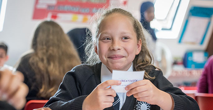 Young student is sat at a classroom table with a card reading 'creativity' in their hand.