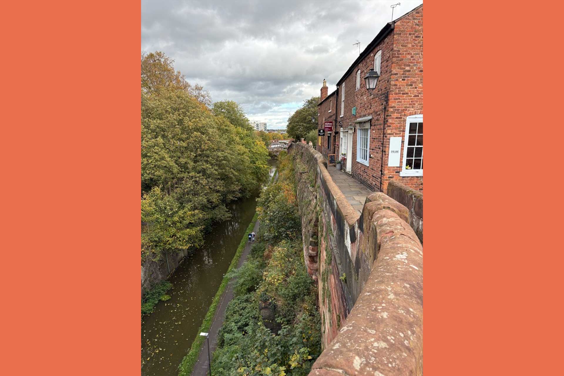 A view of an ancient wall, with a canal outside the wall and trees, with redrick houses inside the wall