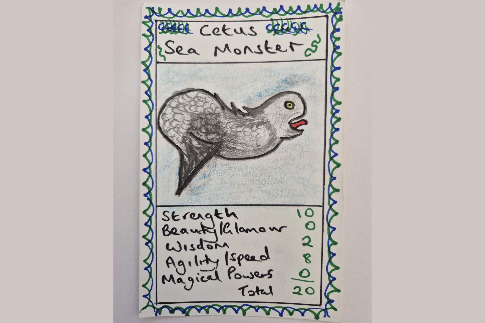 A hand drawn playing card on a piece of paper. There is a drawing of a creature, perhaps mythical. At the top reads Cetus Sea Monster, underneath is a drawing of the creature, then statistics about the creature: Strength - 10, Beauty-glamour - 0, Wisdom - 2, Agility/speed - 8, Magical powers - 0, Total - 20.
