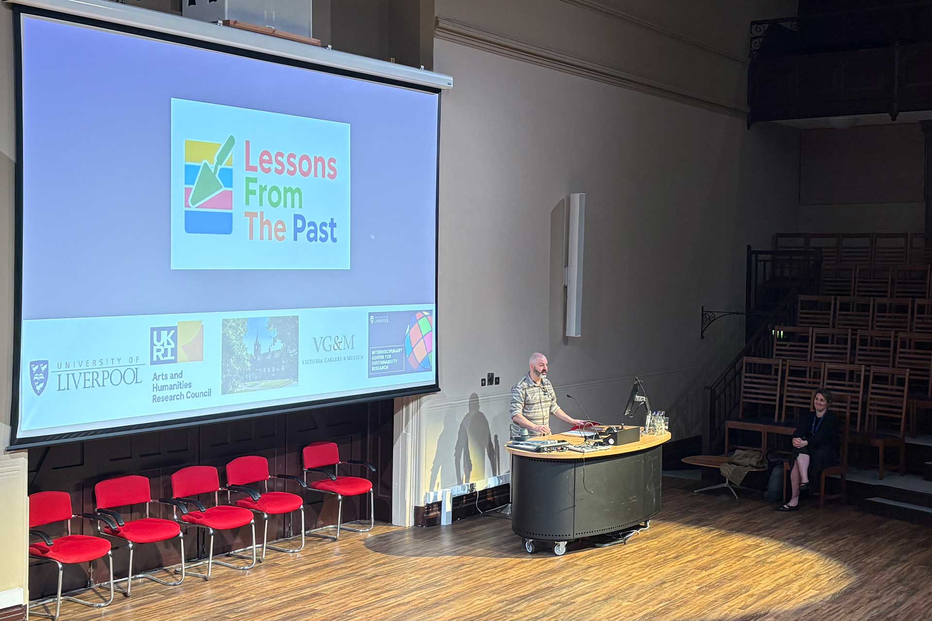 A man stands a the front of a lecture hall in front of a presentation screen that reads 'Lessons of the past'