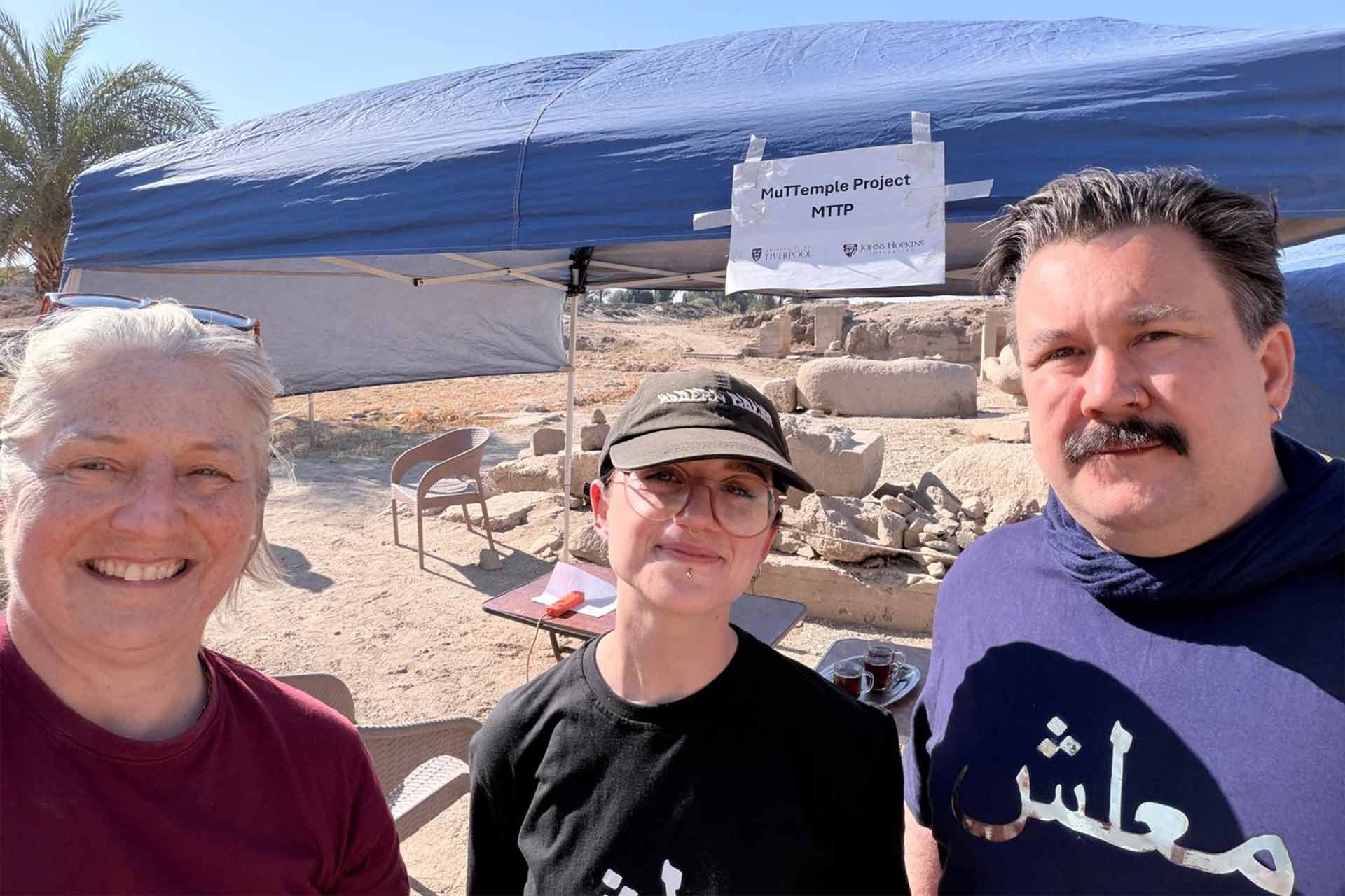 Three people standing and smiling in front of a outside covering, with an A4 piece of paper attached that reads 'Muttemple Project'