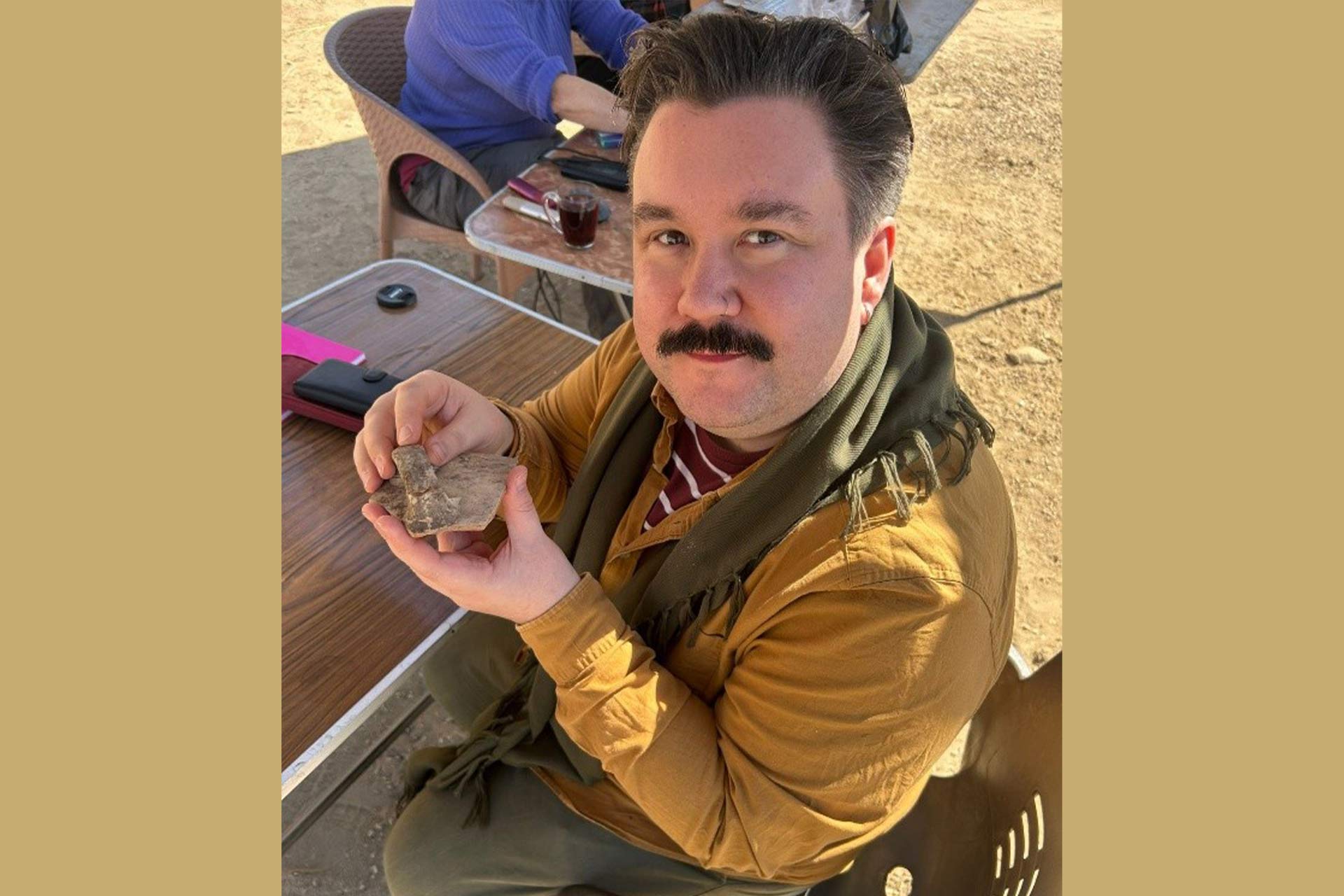 A man sitting outside in a sandy dry area, holding an old item that has been excavated from an Archaeological site.