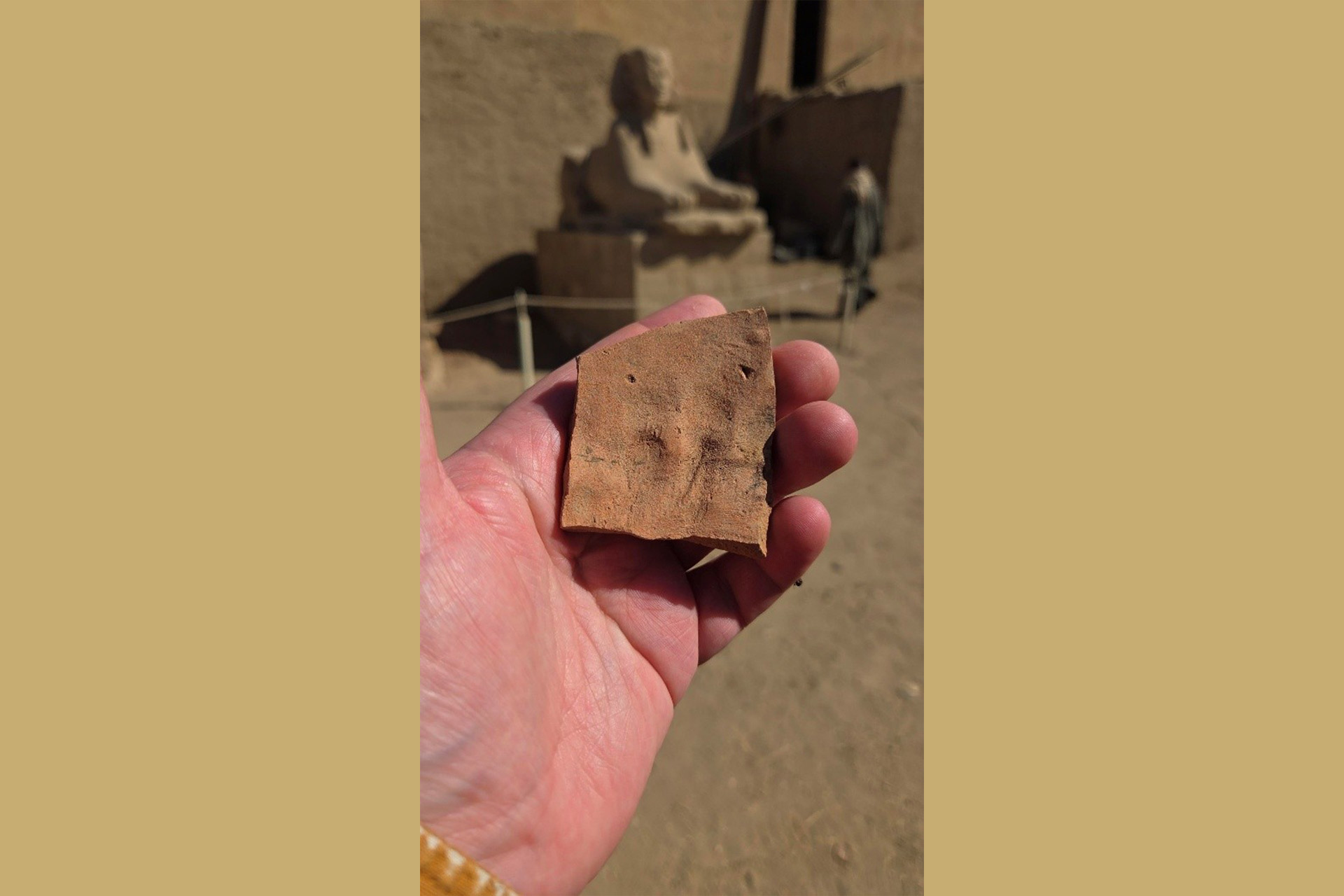 A hand holding a piece of an item, perhaps a broken piece of pottery. An Egyptian statue is in the background.