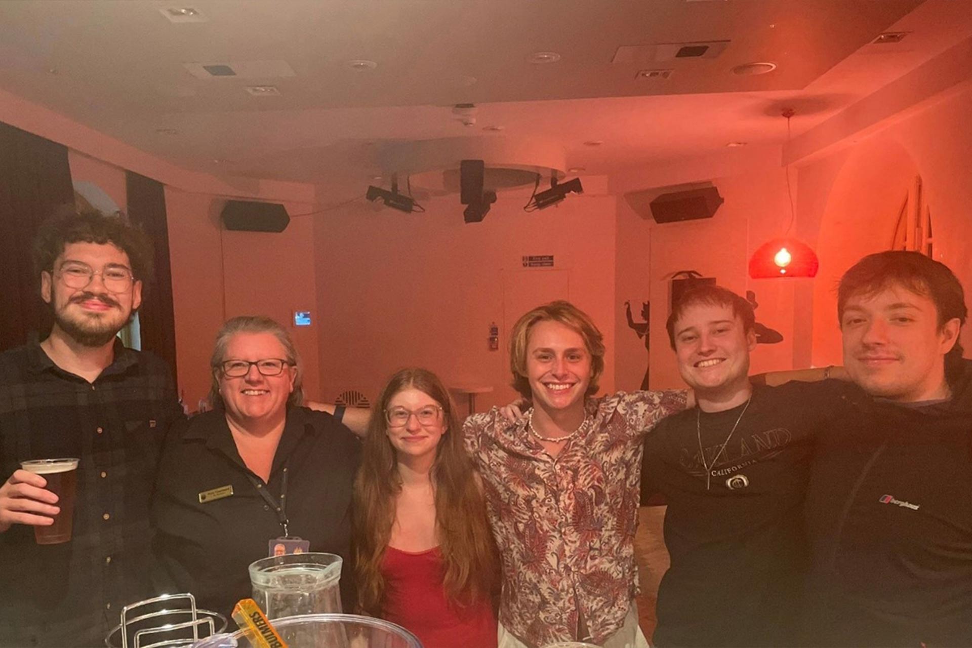A group of 6 people standing together and smiling inside a room in the evening with dimly lit lights.