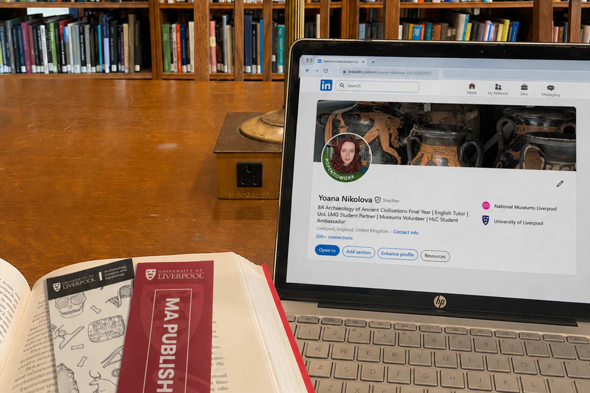 A laptop on a wooden library desk displays a LinkedIn profile page, with a book open beside it and tall wooden bookshelves filled with books in the background.