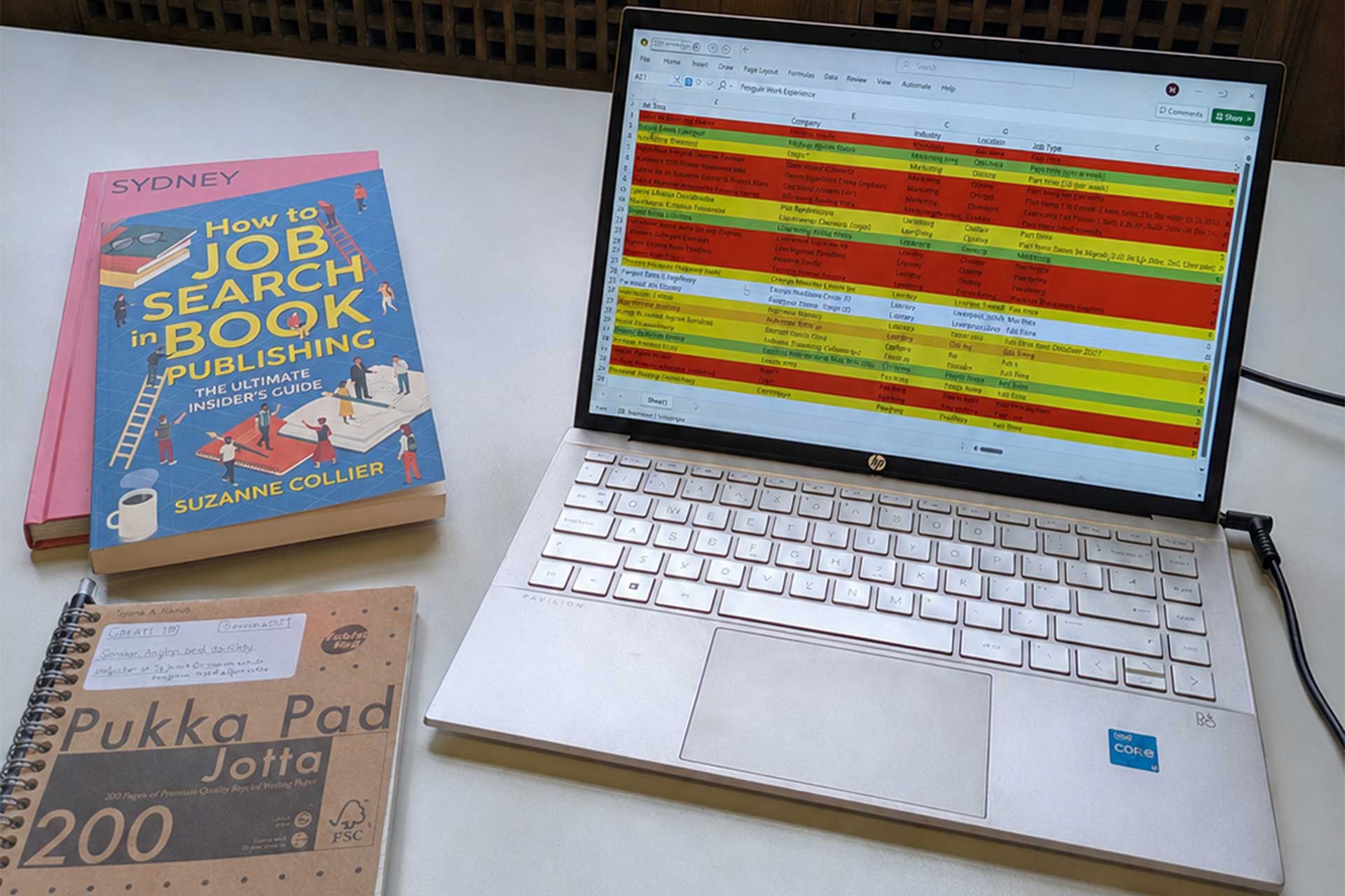 A laptop on a white desk shows a colour coded spreadsheet, next to notebooks and a book titled How to Job Search in Book Publishing.