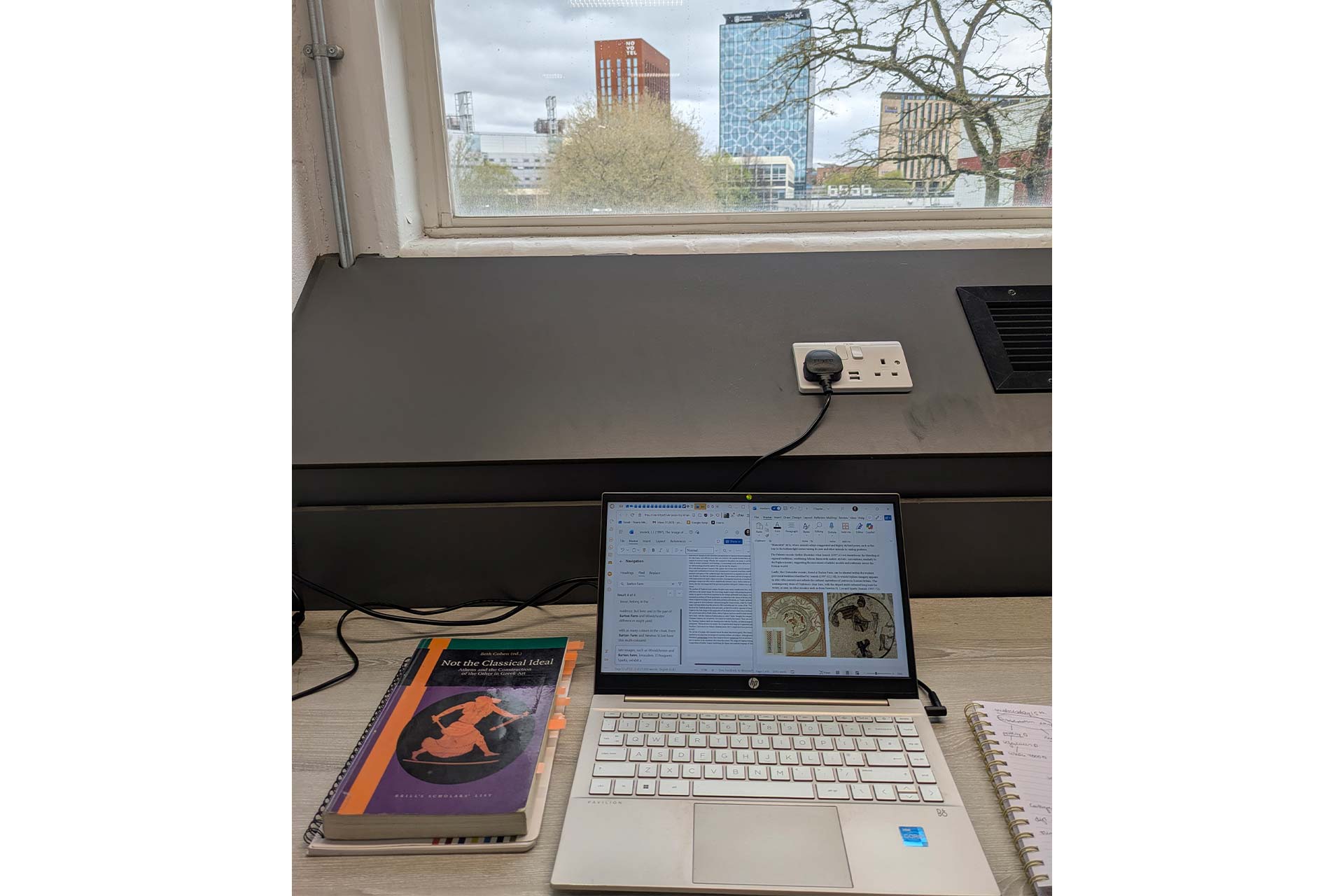 A desk setup with a laptop open on an academic document sits beneath a window looking out onto modern city buildings, with books and notebooks arranged beside the laptop.