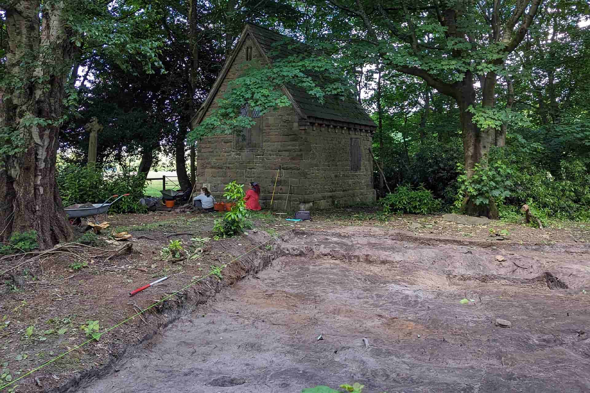 An old chapel in the middle of a wooded areas, with an excavation site in the foreground.