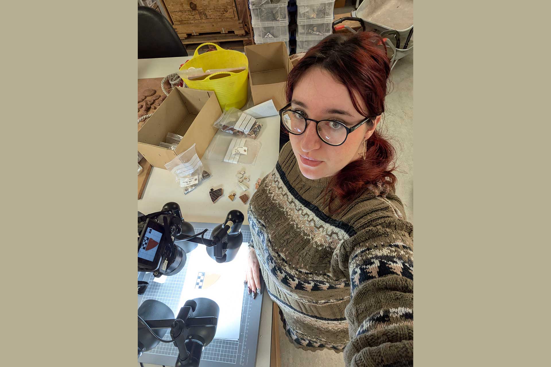Yoana is a female student with long red hair and glasses, she stands by a table with artefacts on it and a bright light to photograph them.