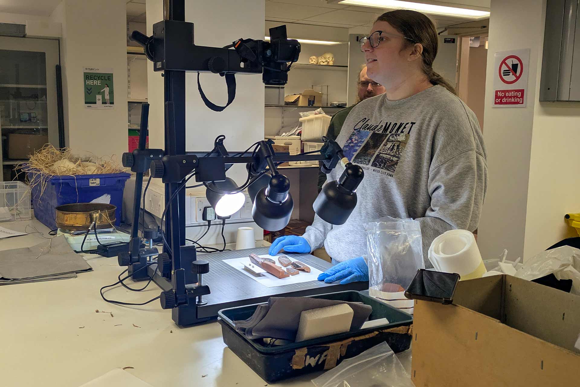 A student in a grey sweatshirt and blue disposable gloves photographing artefacts under bright lights indide.