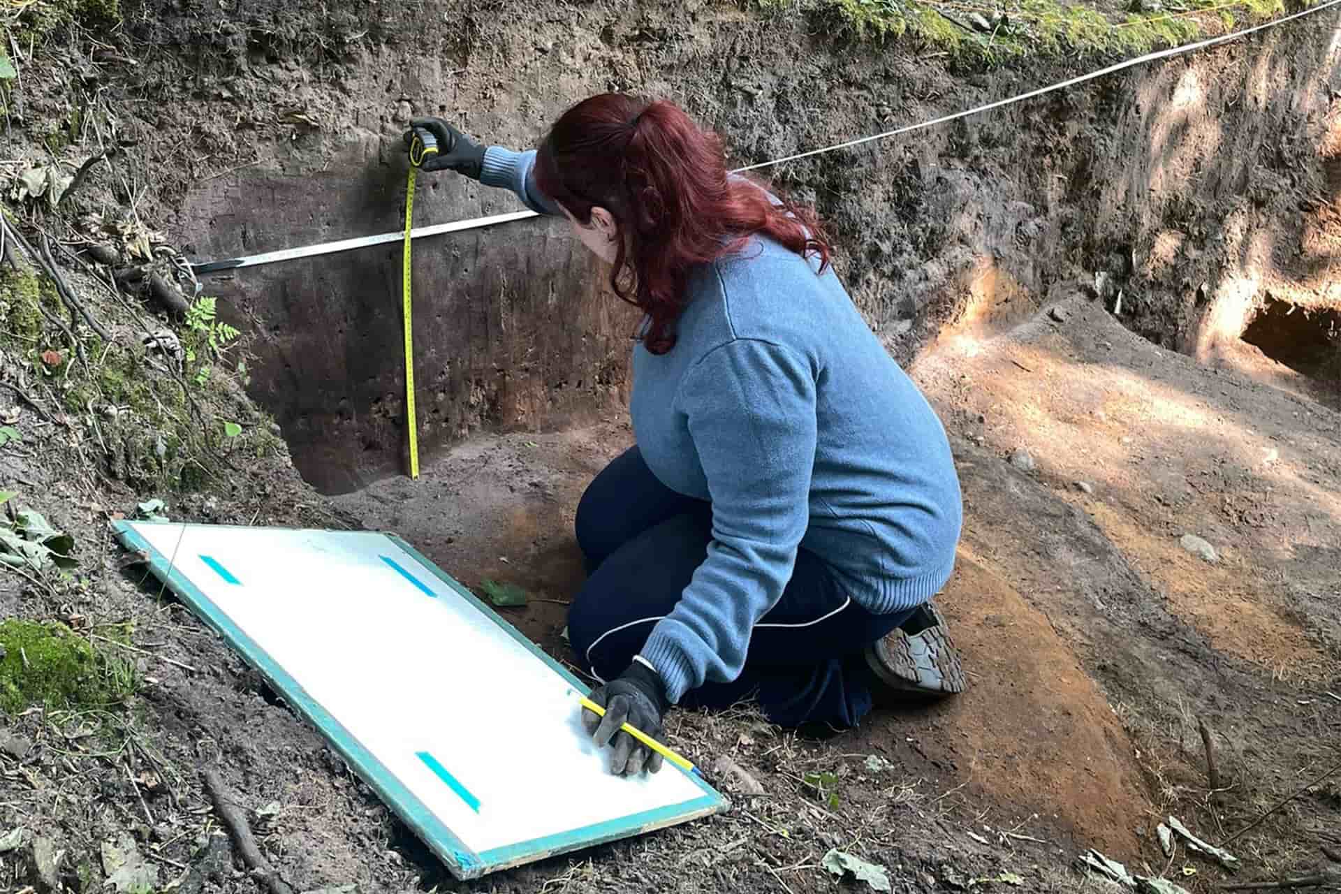 A student kneeling inside a trench, wearing gloves and measuring the trench with rulers.