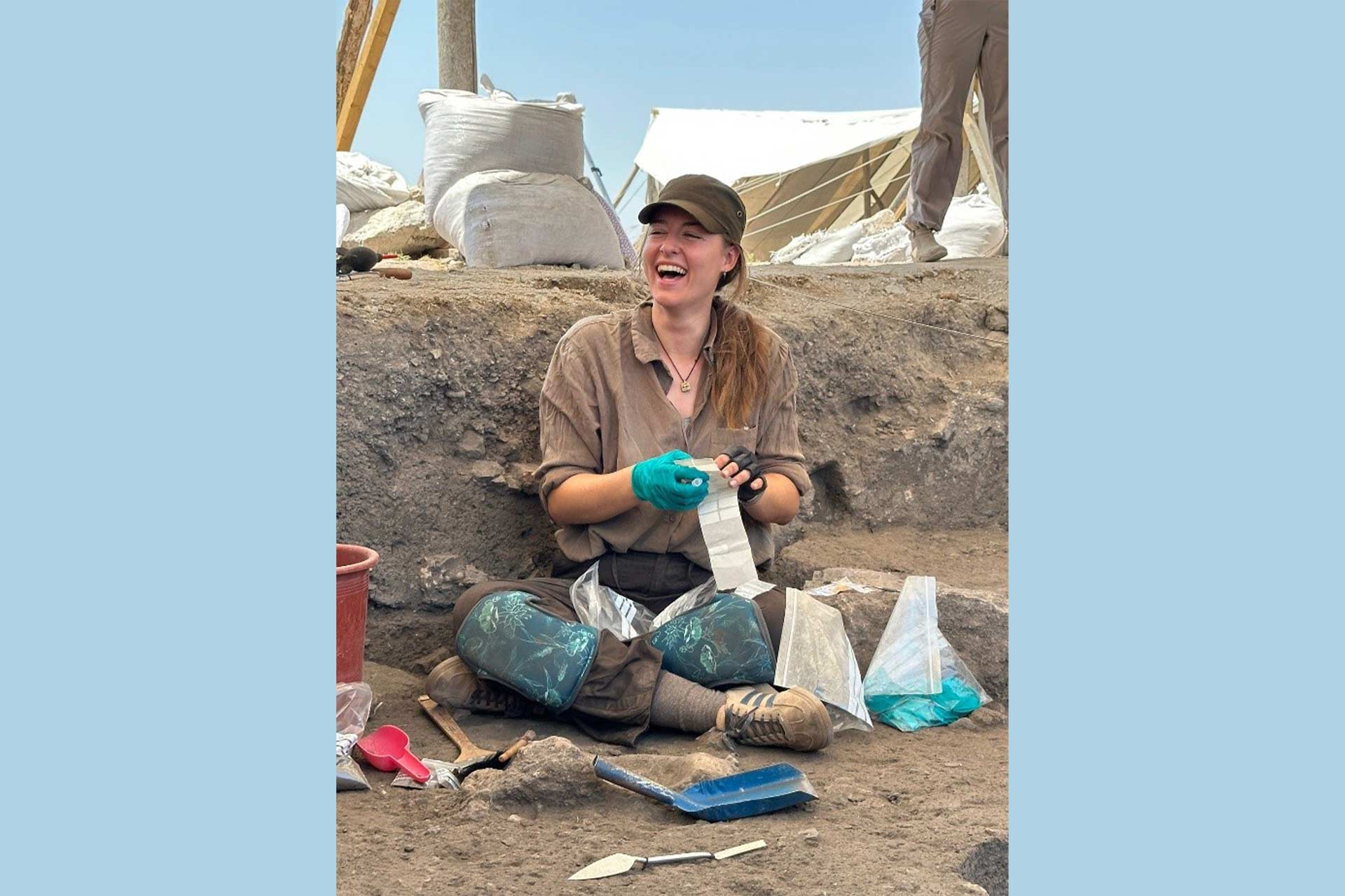 A woman sat outside on dry rocky sandy ground smiling and wearing gloves bagging up Archaeological material.