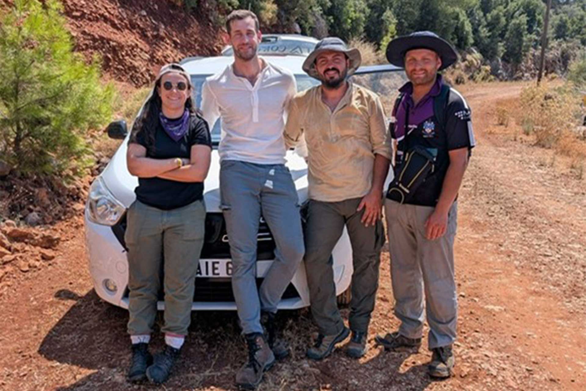 Four people standing together outside in front of a white car.