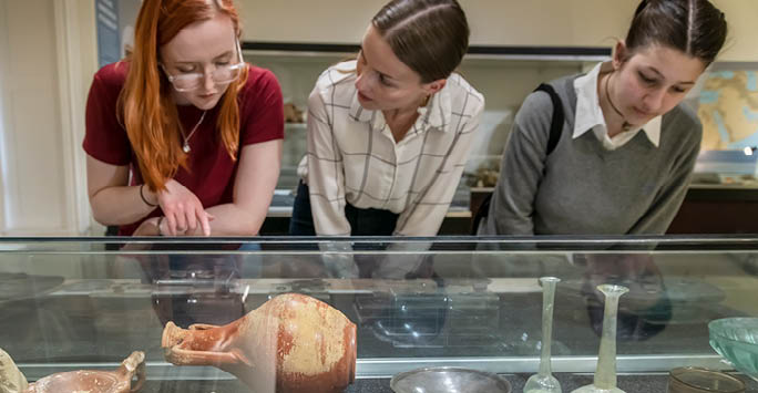 Three students look and point at a cabinet of ancient artifacts