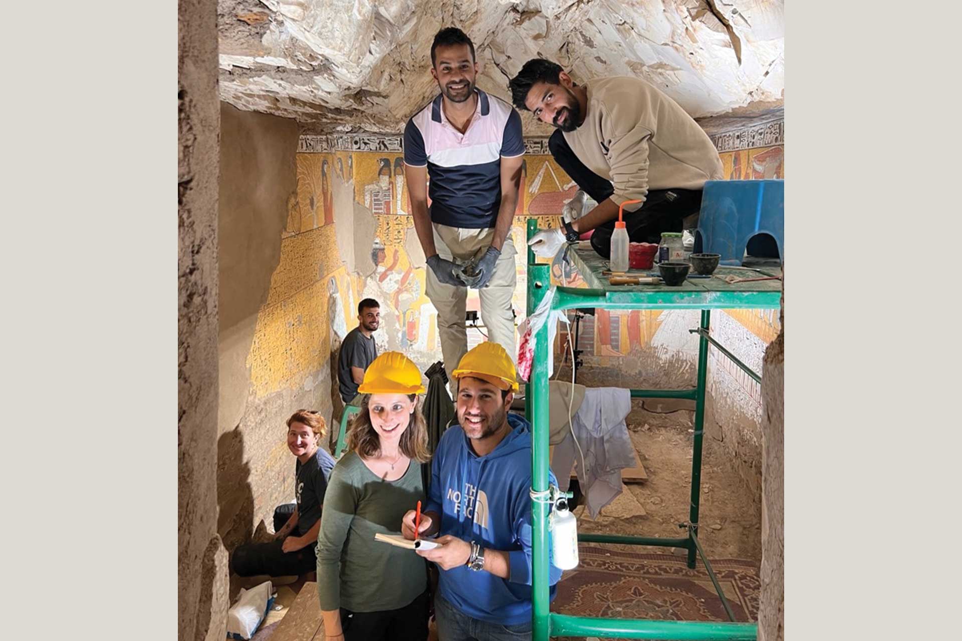 A group of people, some wearing hardhats inside a tomb, perhaps a cave. Egyptian Hieroglyphs are on the walls behind them
