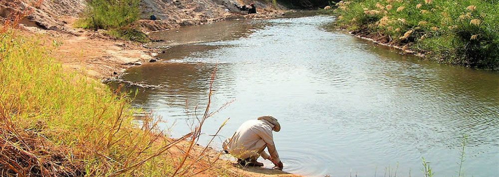 Male archaeologist wearing a beige shirt and wide brimmed hat leaning over looking into a river. The river is located at Kalambo Falls, Zambia