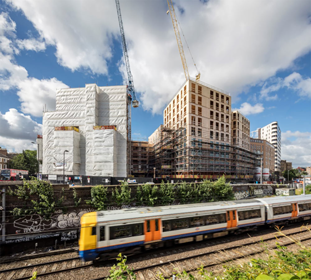 Two buildings, one on the right using cross laminated timber construction, a train is passing in front of the construction site