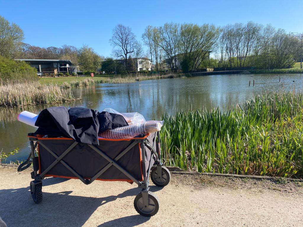 A wheeled trolly in front of a lake, reads and bullrushes surround the lake, on the other side trees obscure some 2 story modern buildings.