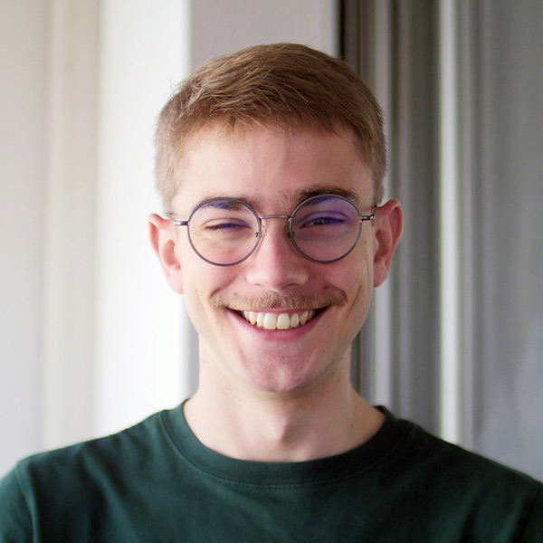 Head and Shoulders of a young white man with short cropped ginger hair and moustache. He is wearing round glasses