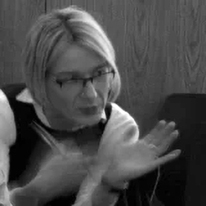 White woman with short blonde bobbed hair and wearing glasses. She is gesticulating with her hands