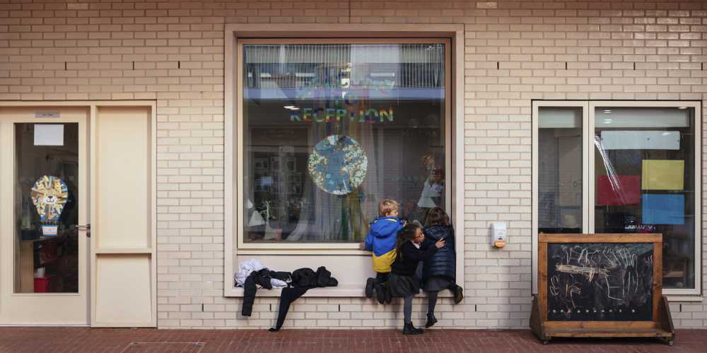 Three children looking in through an internal window set in a brick wall. on the window is a paper cutout world and a rainbow coloured text reading 