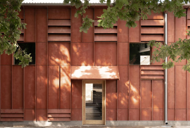 Rust coloured building with prominent vertical ribs and vents, framed by foliage