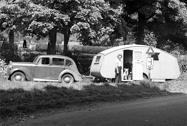 A vintage car and caravan parked by the roadside. A white woman is standing by the open door of the caravan Photo credit, Museum of English Rural Life, University of Reading pulled over in a lay-by with trees behind. A woman stands by the door and a dog looks out from inside the caravan.