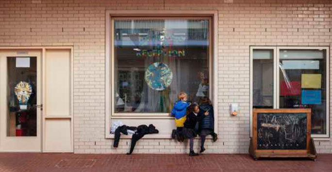 Three children looking in through an internal window set in a brick wall. on the window is a paper cutout world and a rainbow coloured text reading 