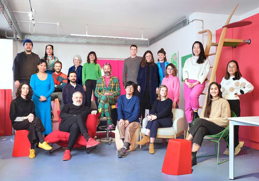 Mixed group of people in two rows some sitting some standing. Geometric furniture.