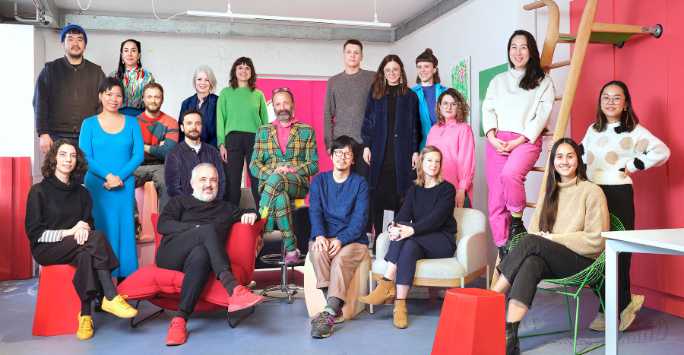 Mixed group of people in two rows some sitting some standing. Geometric furniture.