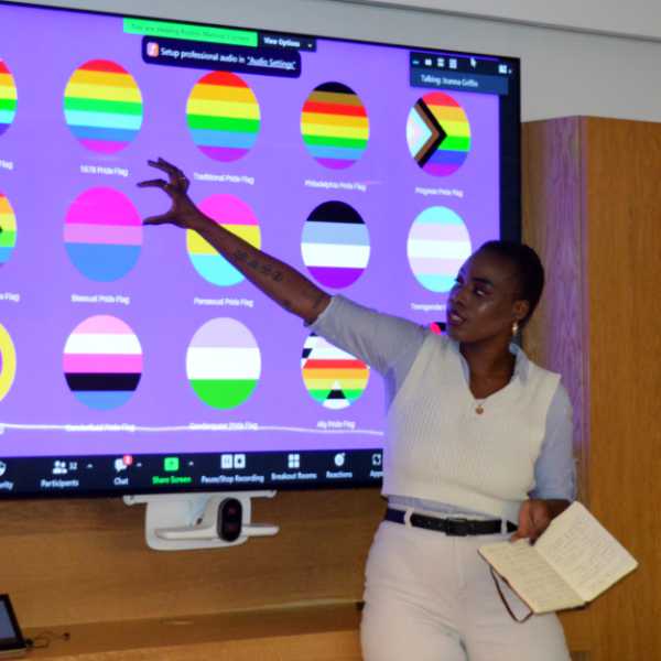 Black woman in jeans and shirt holding an open notebook in their left hand, pointing at slide with their right hand showing different LGBTQI+ pride flags.