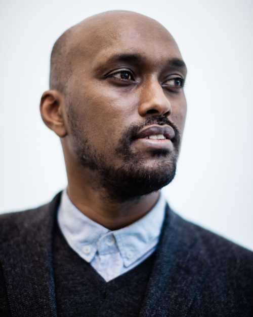 Young black male with shaven head, trimmed beard and buttoned up shirt.