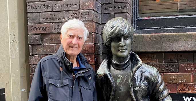 Middle aged man with short whitye hair standing next to a bronze sculpture of John Lennon.