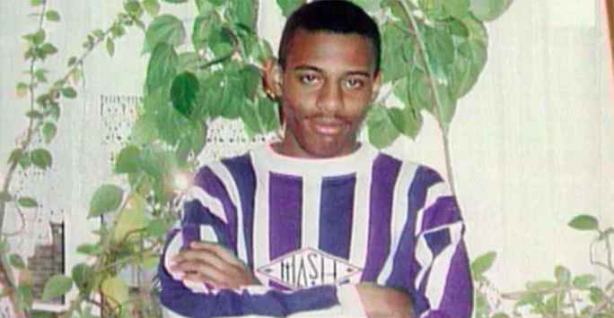 Young black man, smiling at the camera wearing a stripped jumper. He is standing in front of a wall and houseplants.